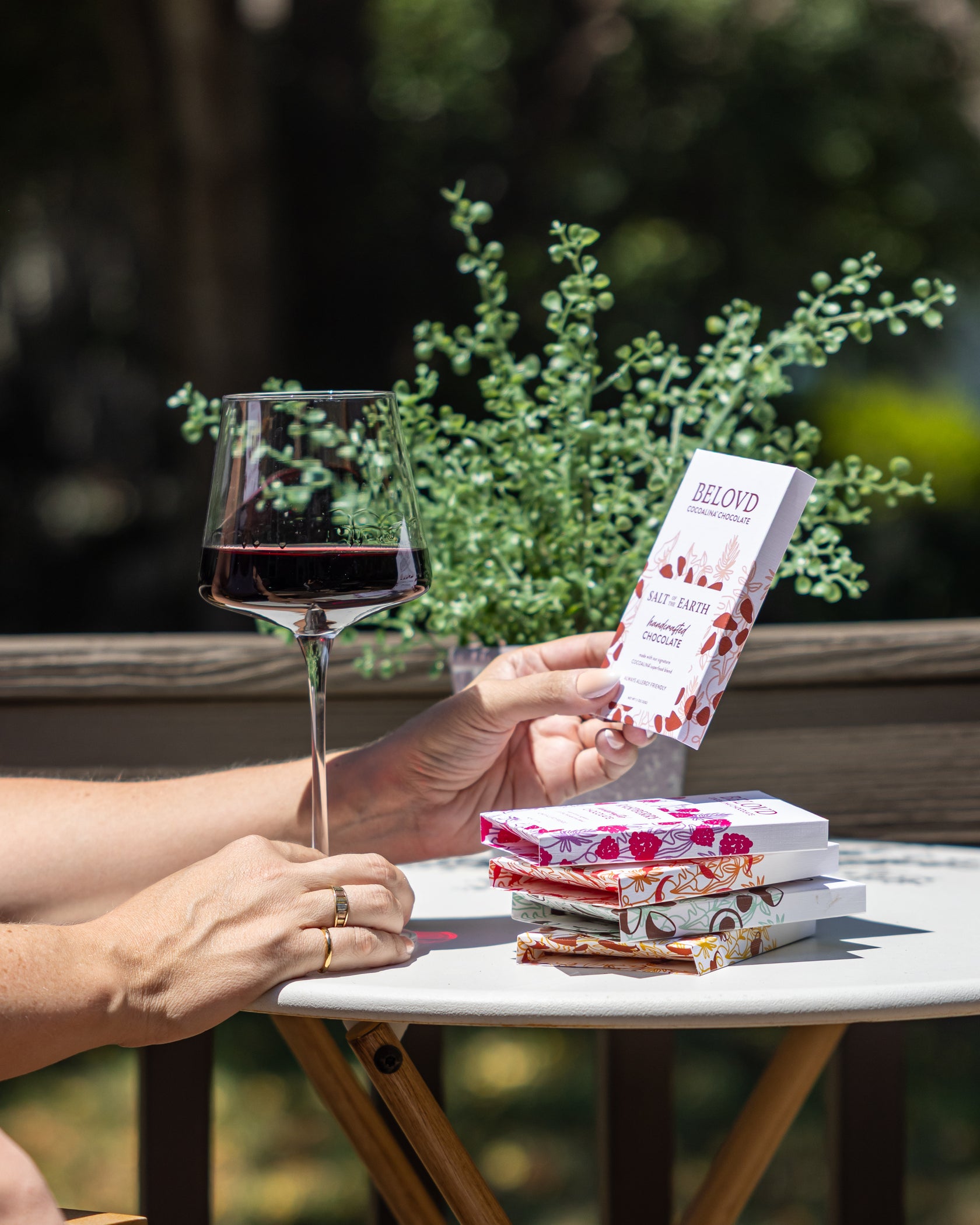 Person holding a glass of red wine and a package of Beloved Period Products on a table outdoors.
