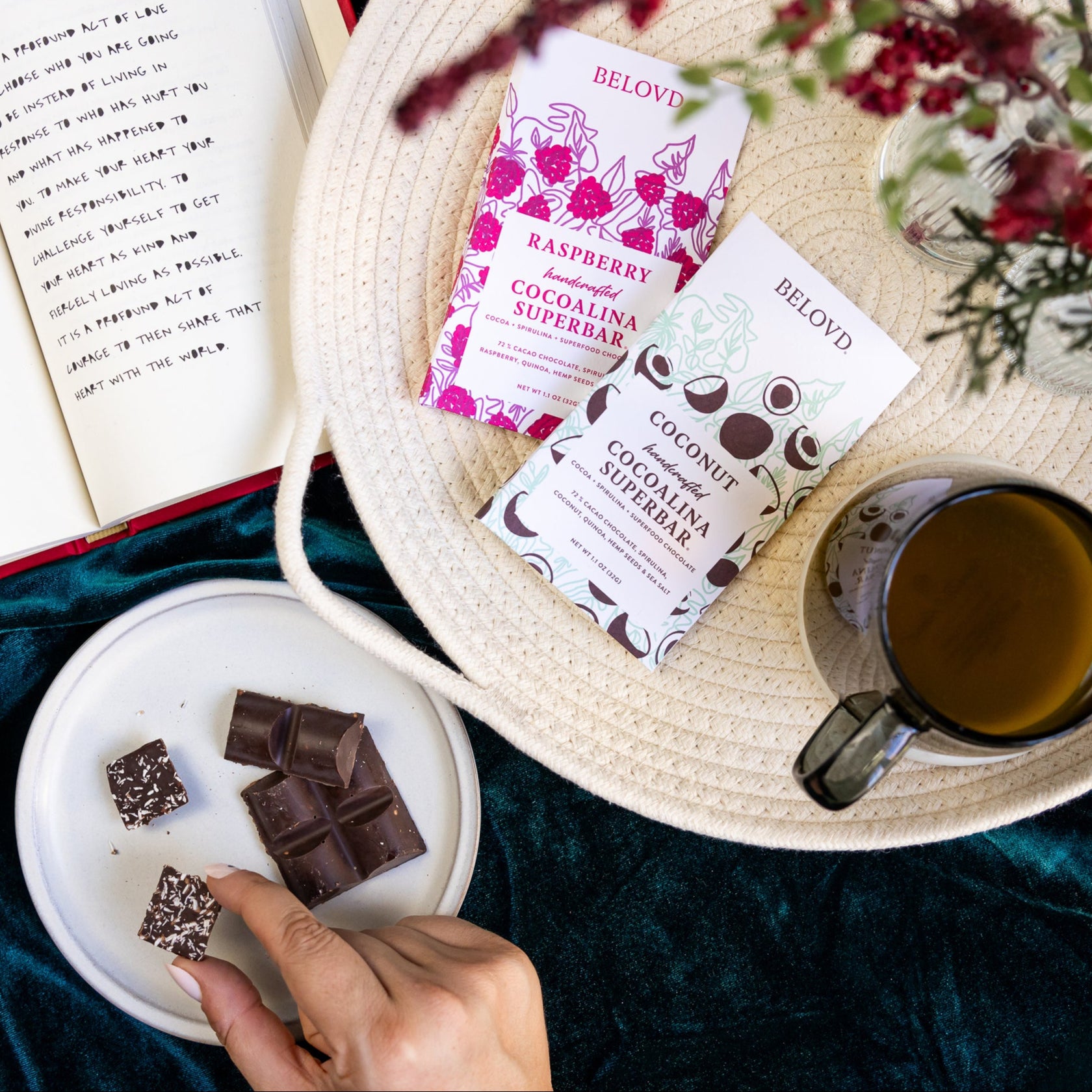 Woman's hand picking up a square of Belovd Coconut chocolate next to a tray of chocolate bars and tea.