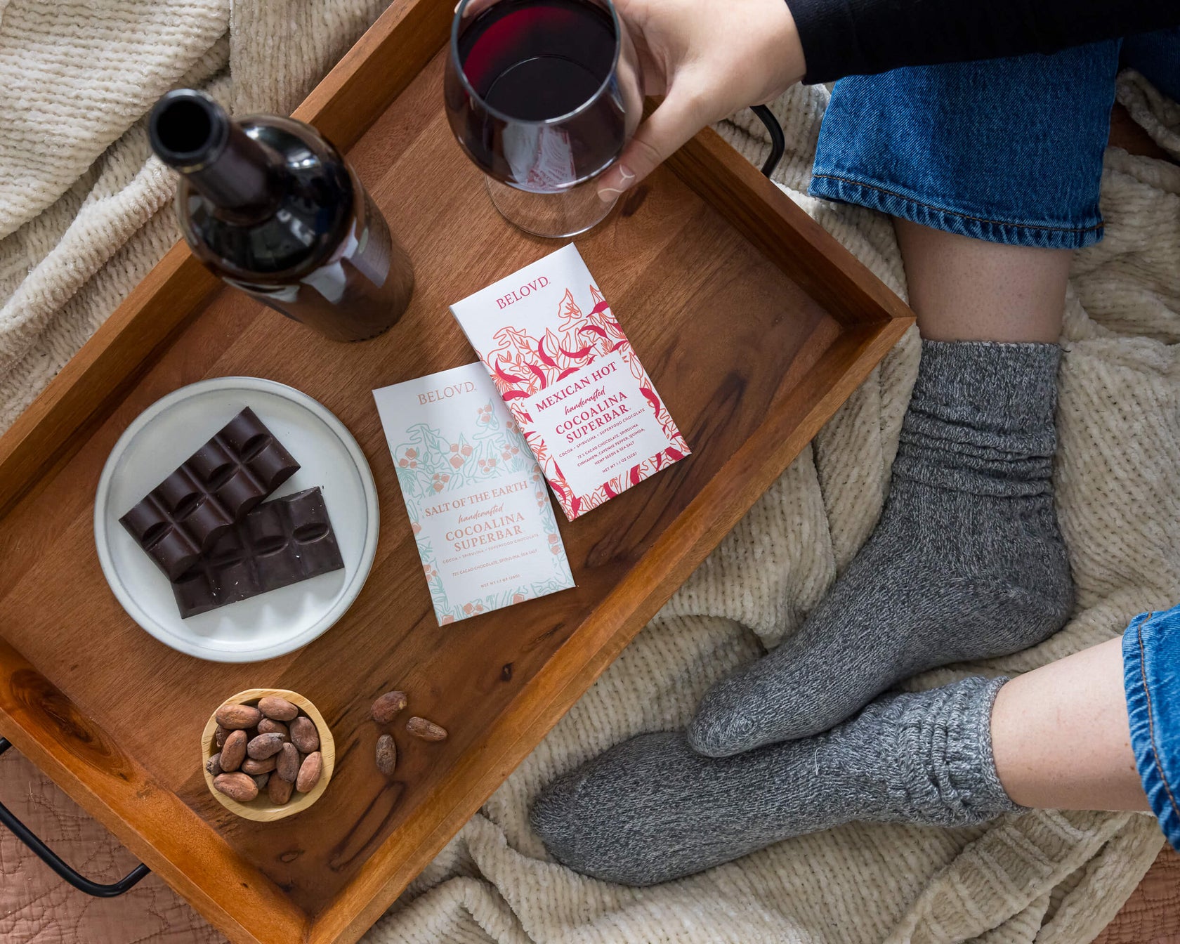 Person holding a glass of red wine with a wooden tray featuring chocolate, a bottle, and cards on a cozy blanket.