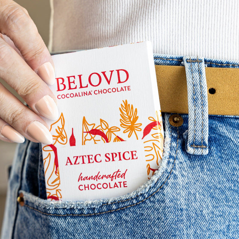 Hand holding a Belovd Chocolate Aztec Spice bar in front of a person wearing blue jeans.
